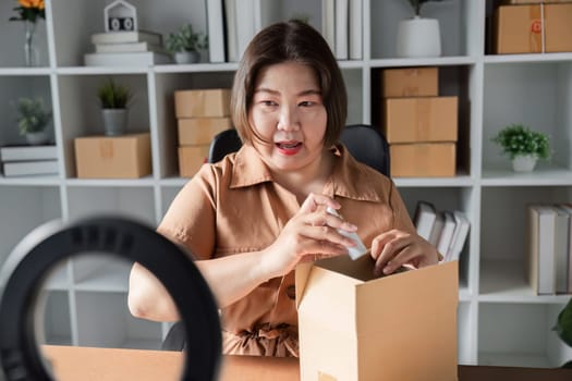 A young woman excitedly pulls out a skincare product from a box, engaging with her audience during a livestream sales session in her well-organized home office.