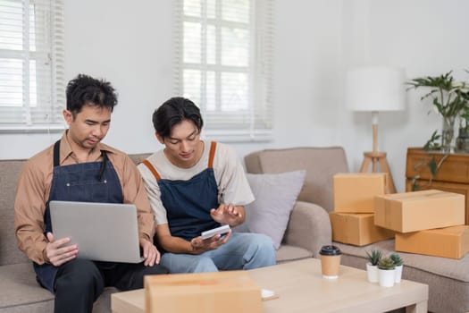Two men focused on reviewing their sales performance using a laptop and smartphone, surrounded by packed boxes in their modern and cozy space.