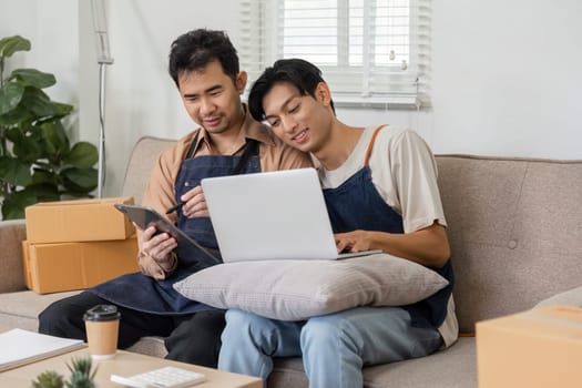 A happy LGBT couple working together on their online business, sharing insights on a laptop and tablet, surrounded by packing boxes in their cozy living room.
