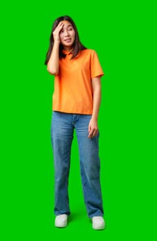 A young woman stands in a bright orange shirt and blue jeans, showing a confused expression. She touches her head, surrounded by a vibrant green backdrop, indicating a playful mood.