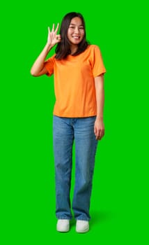 A young woman wearing an orange shirt and blue jeans stands confidently on a bright green background. She smiles and makes a hand gesture indicating everything is okay.