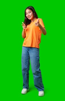 A young woman stands confidently while playfully pointing fingers at the camera, wearing an orange shirt, blue jeans, and white sneakers. The vibrant green backdrop enhances her lively expression.