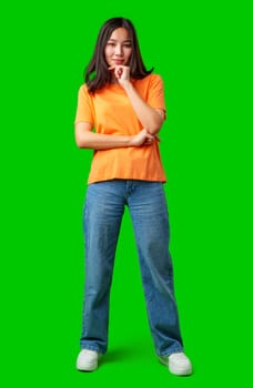A young woman stands with her arms crossed, wearing a bright orange shirt and blue jeans. She has a thoughtful expression while against a vibrant green background.