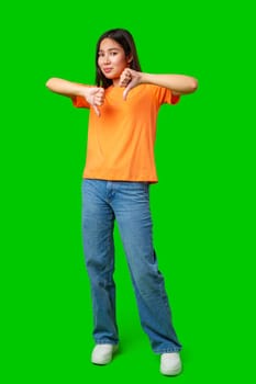 A young woman stands with a thumbs down gesture, wearing an orange shirt and jeans. Her expression conveys disapproval and is highlighted by the vivid green backdrop, creating a striking contrast.