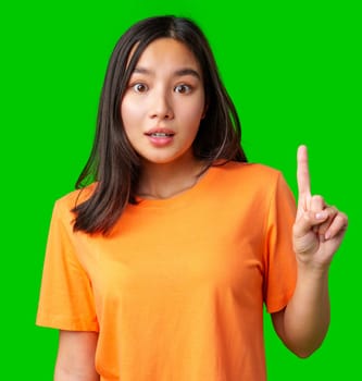 A young woman wearing an orange shirt looks surprised and is raising her index finger. The bright green background adds a lively contrast to her expression, emphasizing her reaction.