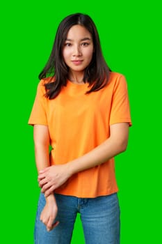 A young woman stands confidently with her arms crossed while wearing an orange shirt and blue jeans. The bright green background enhances her playful look and adds energy to the shot.