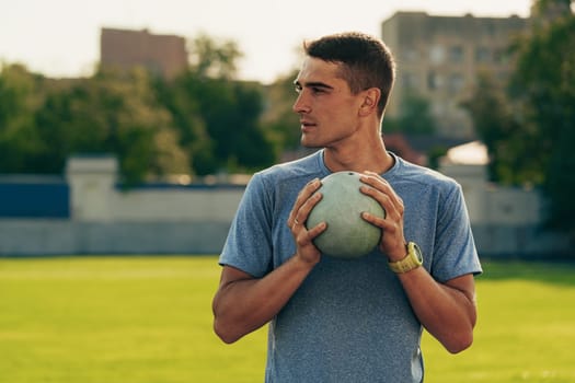 Focused on his technique, a young athlete holds a shot put with determination while standing on a lush outdoor track field. The sun shines brightly in the afternoon sky.