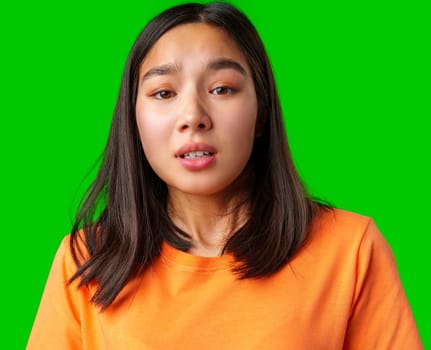 A young woman in an orange shirt is standing against a bright green backdrop, showcasing a range of emotions with her facial expressions. The vivid colors enhance her look.