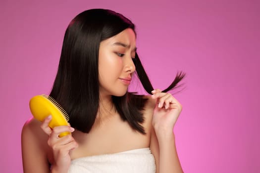 A young woman is engaged in a beauty routine, brushing her straight hair with a yellow comb while looking at her reflection. The vibrant pink background enhances the atmosphere.