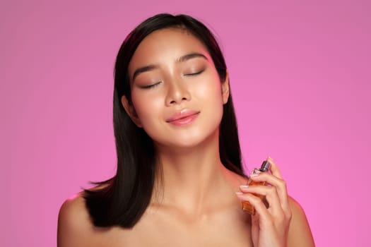A woman enjoys the scent of her perfume while holding the bottle close to her face. She has a serene expression, surrounded by a soft pink backdrop, conveying relaxation and confidence.