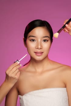 Bright pink backdrop enhances the beauty studio atmosphere as a woman applies makeup, holding a highlighter in one hand and a brush in the other, showcasing a makeup routine.
