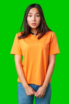 A young woman stands with a surprised expression, wearing an orange shirt and blue jeans. Her hands are held in front, reflecting a moment of shock or disbelief.