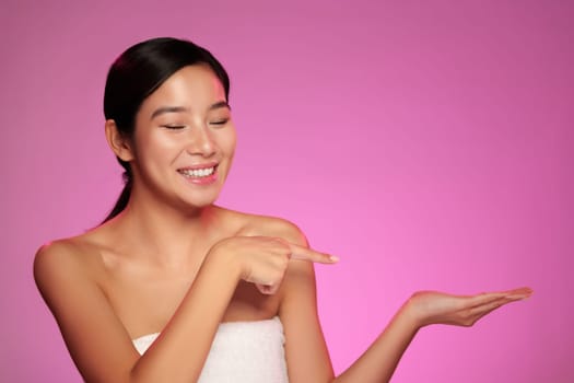 A joyful woman wearing a towel is pointing towards her palm while smiling. The bright pink backdrop enhances her cheerful expression and inviting gesture.
