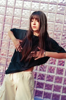 A young woman showcases her dance moves against a textured glass block wall in soft afternoon light, expressing emotion through graceful motions and focused concentration.