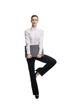 Full body young woman in formal wear carrying laptop and looking at camera while balancing on leg against white background