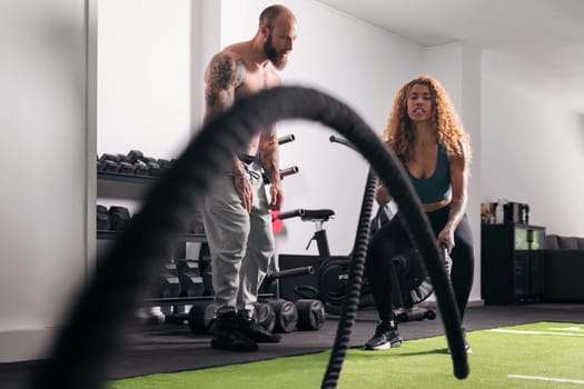 muscular sportswoman doing exercise with rope during functional workout with male buddy at gym, concept of fitness and active lifestyle