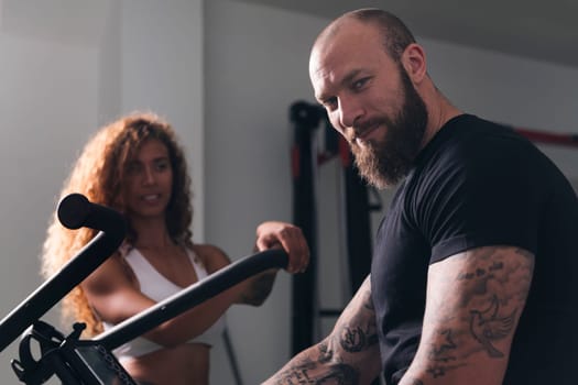 confident muscular bodybuilder with tattoos looking at camera against cheerful sportswoman at gym, concept of fitness and active lifestyle