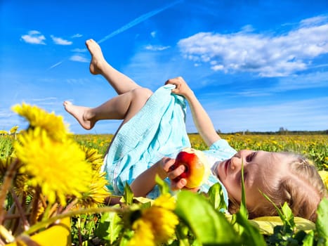 A young girl lies among blooming dandelions, smiling and holding an apple, enjoying a bright sunny day in the countryside.