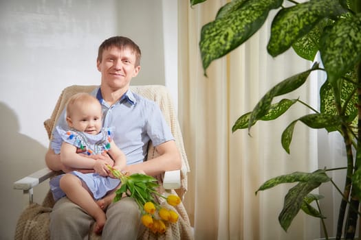 A joyful father sits in an armchair holding his smiling baby daughter, who is wrapped in his embrace while proudly clutching a bouquet of yellow tulips.