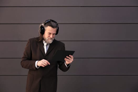 serious mature male with beard in formal suit and wireless headphones using tablet while communicating online against gray wall, concept of middle age business man and technology of communication