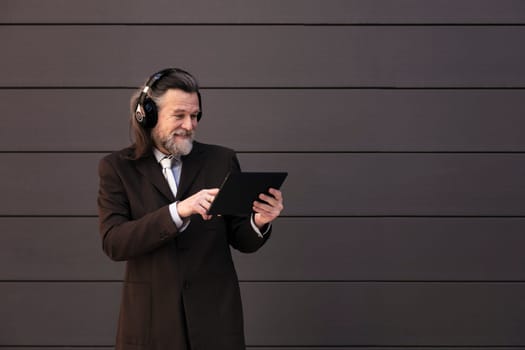 content mature male with beard in formal suit and wireless headphones using tablet while communicating online against gray wall, concept of middle age business man and technology of communication