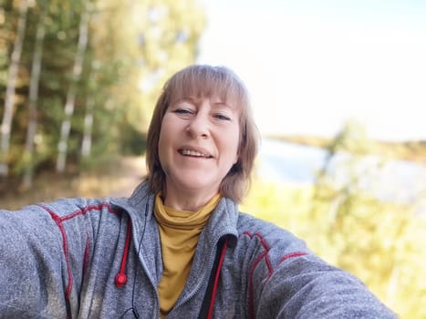 A happy middle-aged woman captures a selfie while on a nature trip in autumn, surrounded by colorful foliage near a lake.