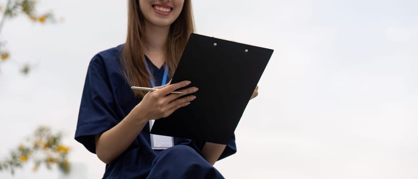 A cheerful female healthcare professional in scrubs holds a clipboard outdoors, emphasizing the importance of relaxation and focus in her medical practice.