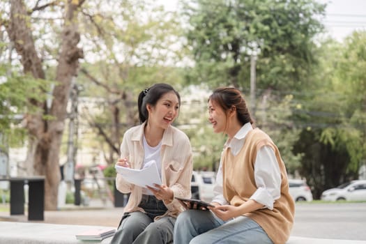 Two diverse female students laughing together while studying, sharing notes and using a tablet in a park-like setting on their university campus.
