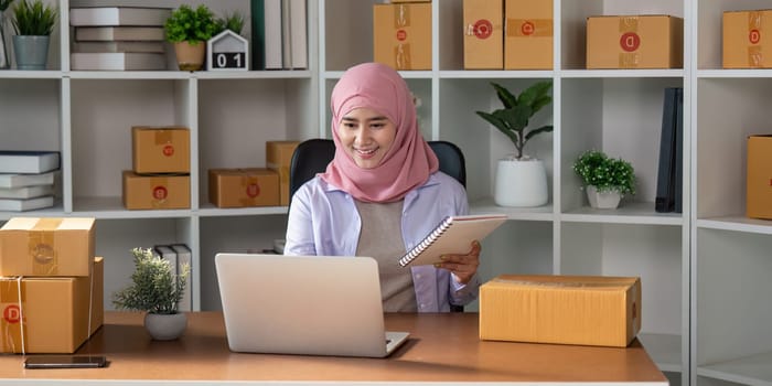 A Muslim woman in a hijab joyfully managing her online sales using a laptop and taking notes in her home office, reflecting her dedication to her business.