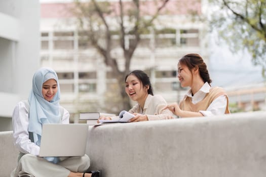 Three diverse female students collaborating outdoors at a university, using a laptop while sharing notes and enjoying their study session together.