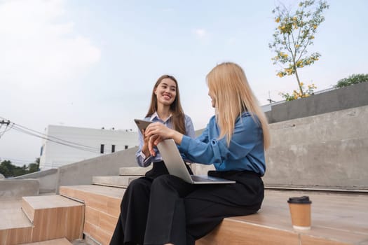Two businesswomen sitting on outdoor steps, collaborating and sharing ideas while using a laptop and a tablet in a professional environment.