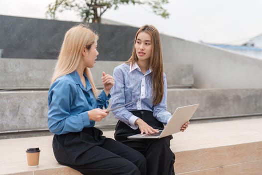 Two diverse women engaged in a collaborative discussion while working on a laptop in an outdoor setting, fostering creativity and teamwork.