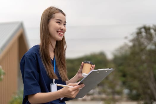 A happy female healthcare professional in scrubs enjoys a moment of rest with a clipboard and coffee outdoors, emphasizing the importance of relaxation in her role.