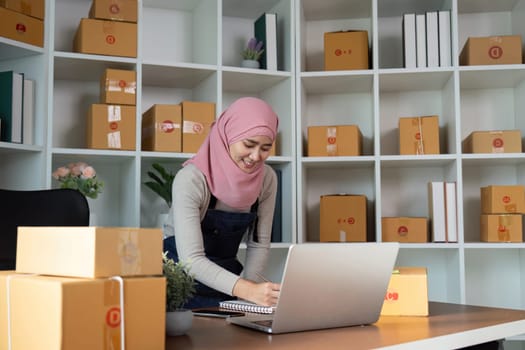 A focused Muslim woman diligently writing notes and managing her online sales in a neatly organized workspace, reflecting her entrepreneurial spirit in e-commerce.