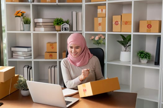 A cheerful Muslim woman engaged in online sales, smiling at her laptop while holding a package, showcasing her enthusiasm for e-commerce in a well-organized workspace.