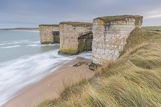 Rocky cliffs rise dramatically from a sandy beach as waves crash against them during cloudy weather.