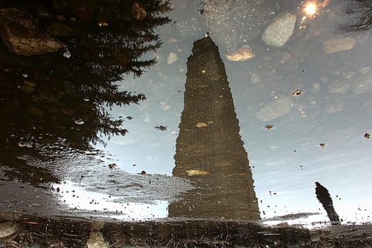 A tall stone tower stands reflected in a puddle, surrounded by scattered rocks and a person walking in the foreground.