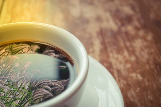 Grass flower (poaceae) reflection on Coffee in white cup on wood table , process in soft orange sun light style