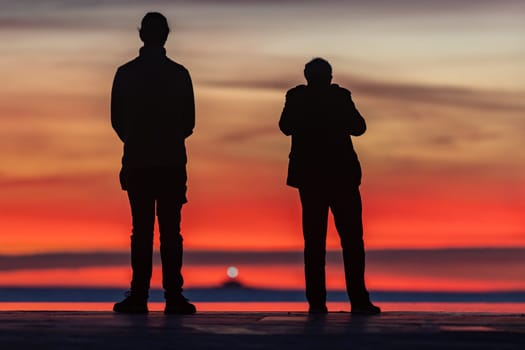 Young couple standing at the seafront and looking at bright red sunset. Mid shot