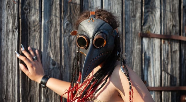 A young nude woman, roped up in a plague raven mask with dreadlocks and piercings, poses near an old wooden fence