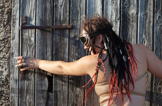 A young nude woman, roped up with dreadlocks and piercings, poses near an old wooden fence