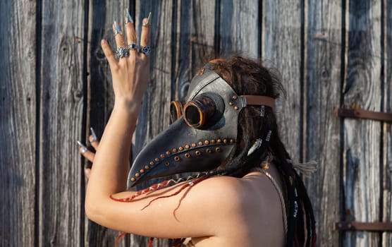 A young nude woman, roped up in a plague raven mask with dreadlocks and piercings, poses near an old wooden fence