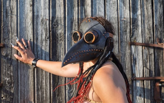 A young nude woman, roped up in a plague raven mask with dreadlocks and piercings, poses near an old wooden fence