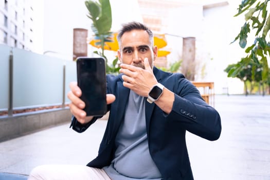 A surprised businessman in a suit covers his mouth while looking at his smartphone. His shocked expression suggests unexpected news or a surprising message in an outdoor urban setting.