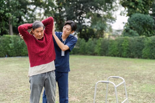 A dedicated health caregiver helps an elderly man put on a sweater while utilizing a walker outdoors in a serene park environment, focusing on rehabilitation and support.