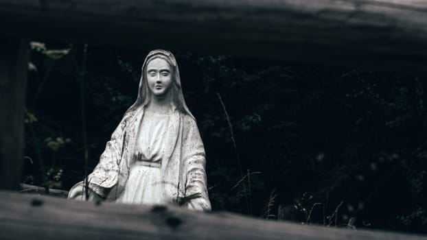 Tucked away in the dense greenery, the statue of the Virgin Mary seems to watch over the forest. The dark woods and somber skies give the scene a mystical quality.