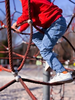 Young caucasian child climbing rope structure at playground in red shirt and jeans.