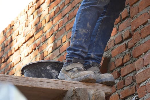Worker balances on a wooden platform while laying bricks in sunlight.