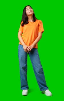 A young woman stands confidently, wearing an orange shirt and blue jeans. She gazes upward, reflecting a curious and contemplative mood in a vibrant green environment.