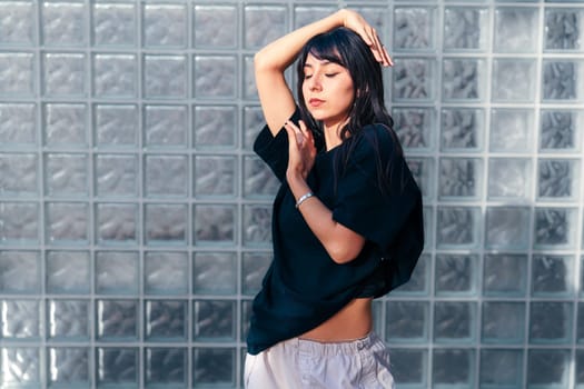 A woman is dancing elegantly against a textured glass block wall. She expresses movement with fluidity, enjoying the sunlight streaming into the space.